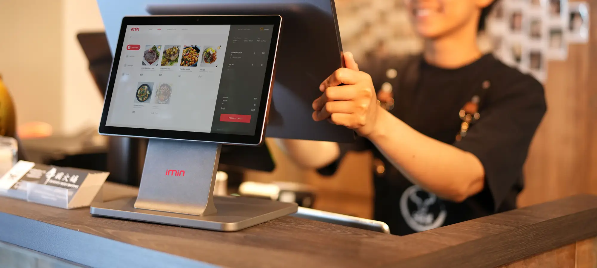 A café worker at a countertop point-of-sale terminal showing a menu of dishes