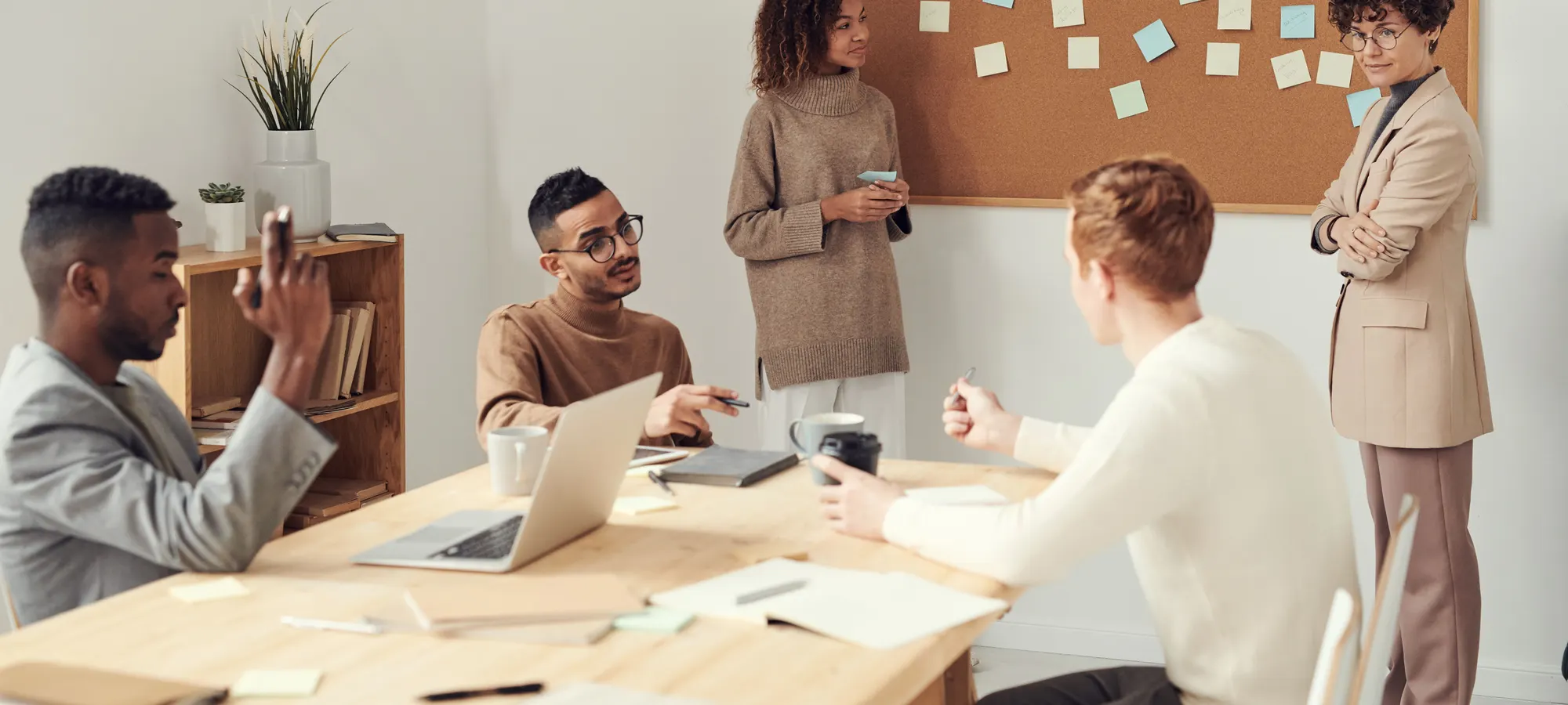 A team standing and seated around a meeting-room table beside a corkboard covered in sticky notes