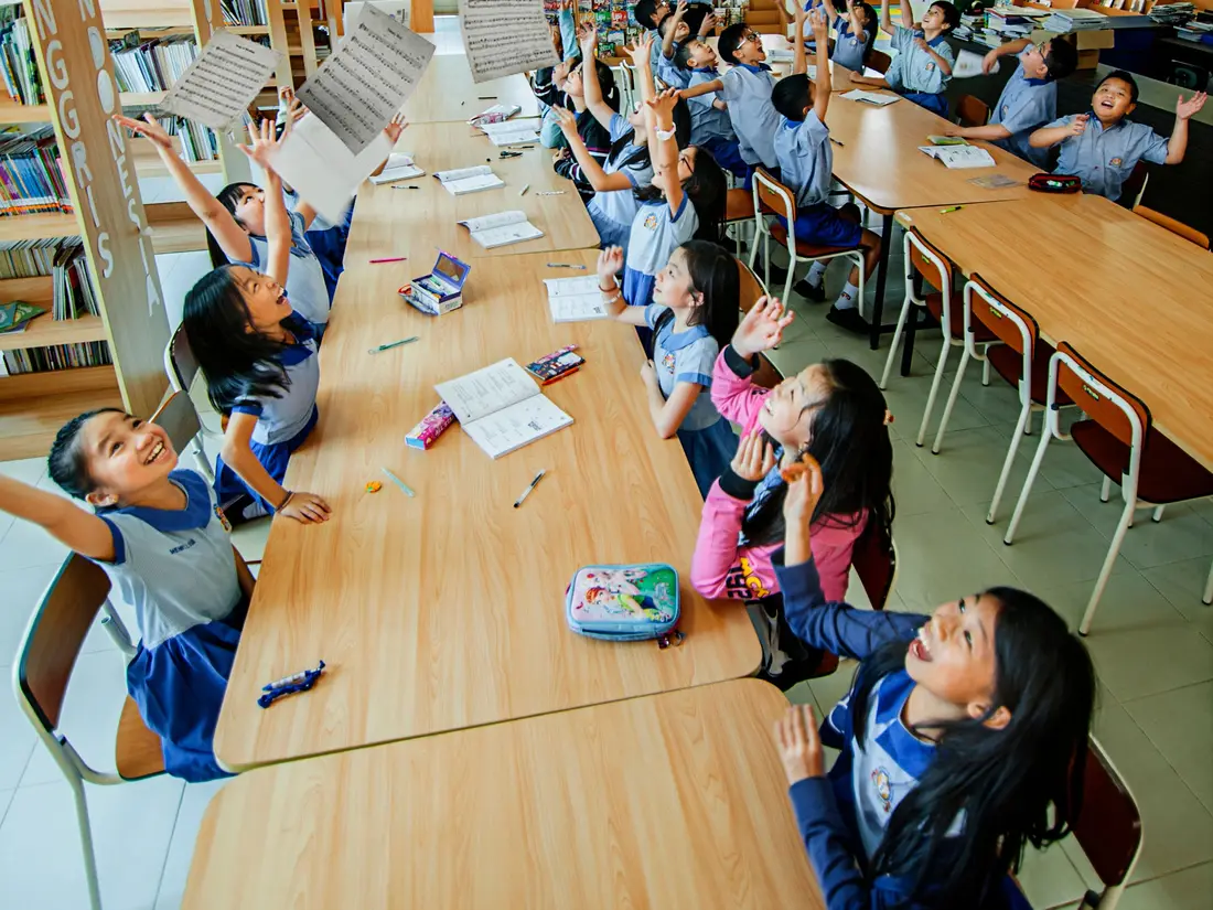 Students in a school library raising their hands among sheets of paper