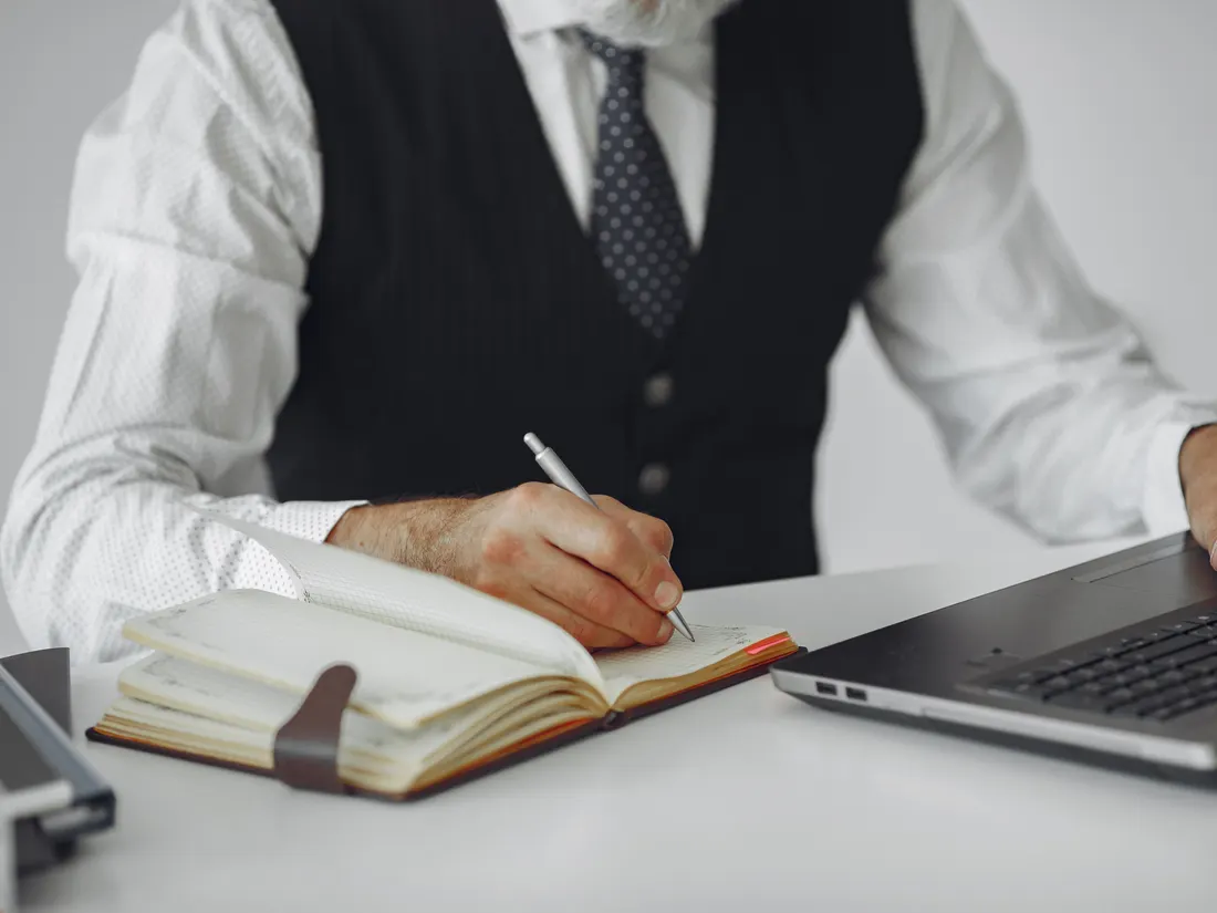 A person in business dress writing in a notebook beside an open laptop