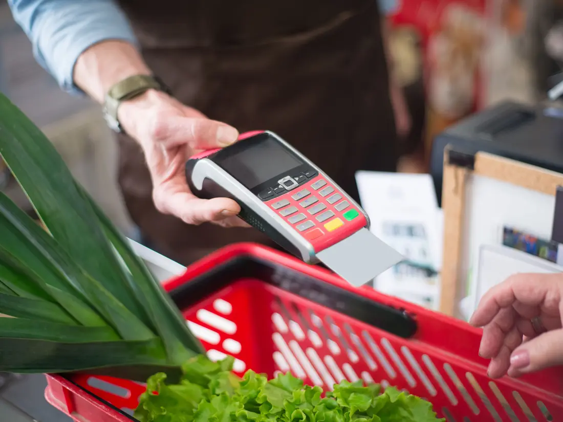 A shopper paying with a handheld card terminal above a red grocery basket
